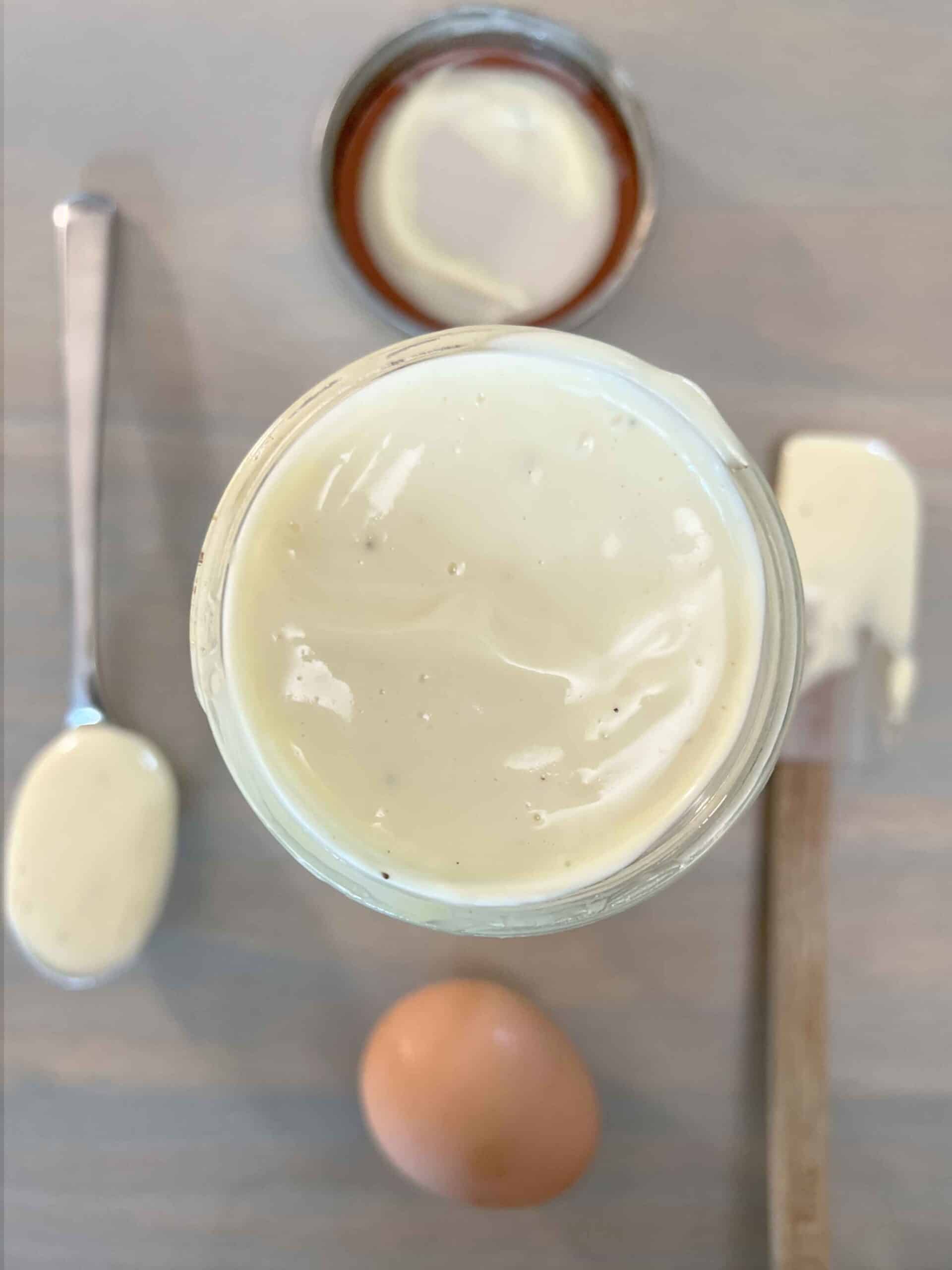 Aerial view of open jar of avocado mayo on countertop arranged with spatula and spoon full of mayo.